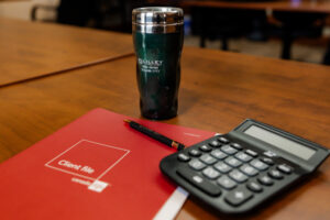 A still life photo on a wooden table showing a 'Client File' in a red folder, a black pen, a calculator, and a green insulated mug. The mug has the 'Lilley Financial' logo and Mike James' name and phone number on it. The background is a blurred office setting with chairs.