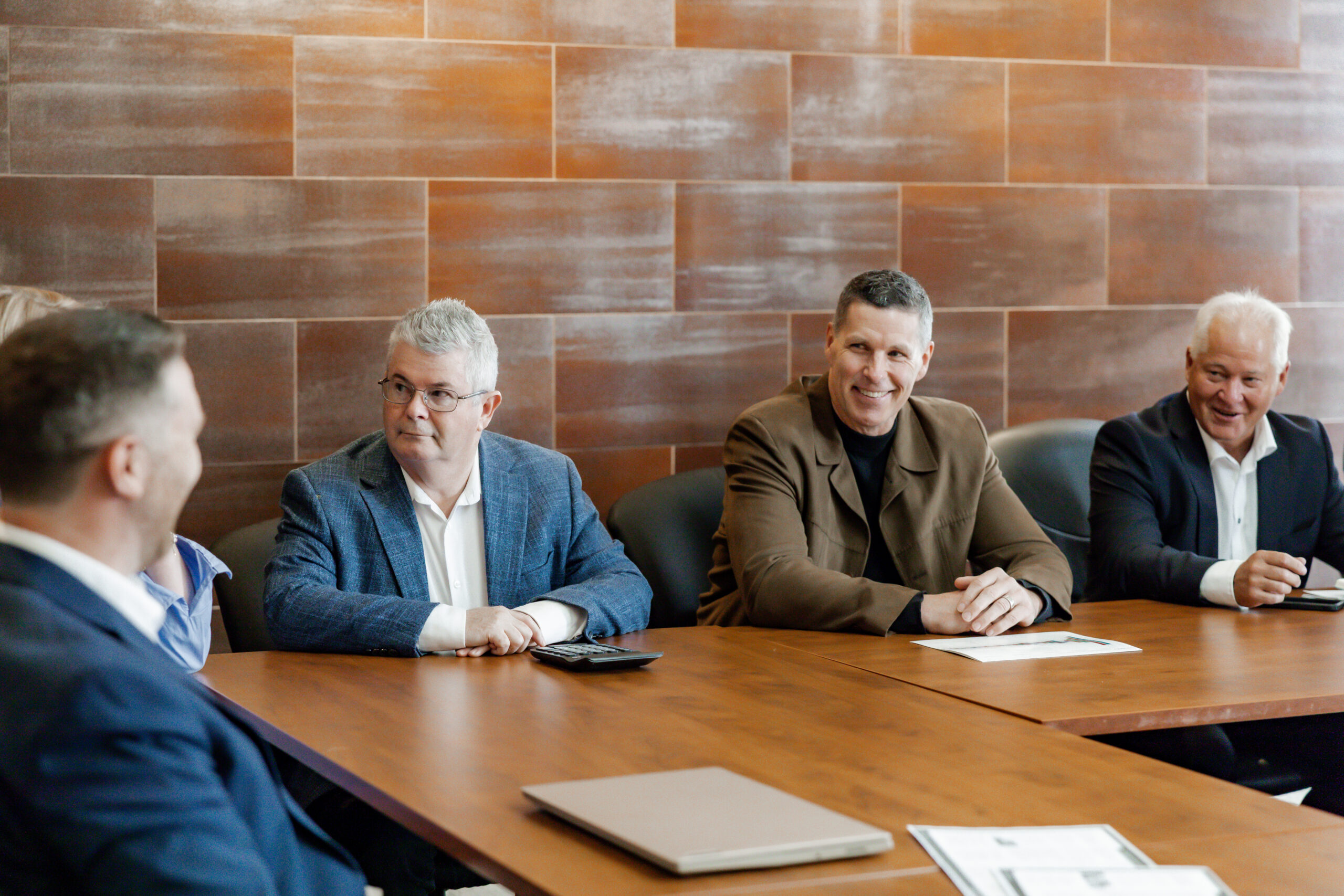 A photo of a business meeting, from left to right: Craig Lilley, Michael Godfrey, and Jim Fockens are seated at a wooden conference table, smiling and looking toward Mike James just out of frame. Craig has a calculator in front of him. The background is a tiled wall.
