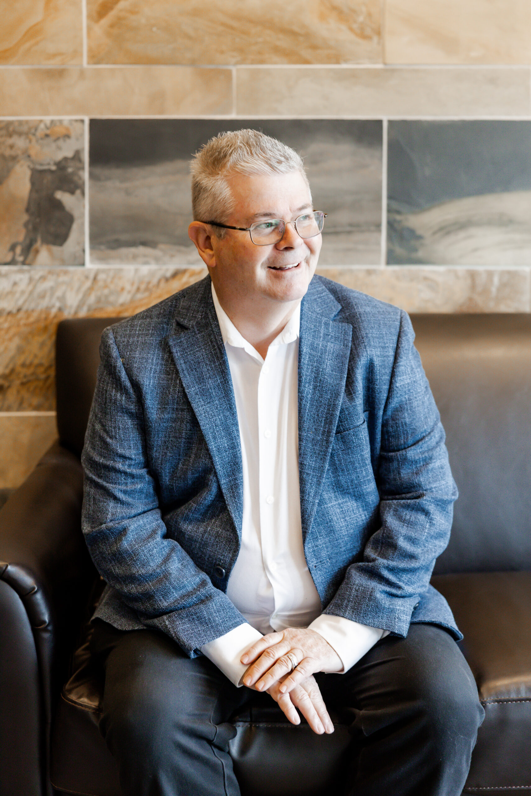 Craig Lilley sits on a black leather couch, smiling and looking to the side. He is wearing a light blue suit jacket and a white dress shirt, with his hands resting in his lap. The background is a tiled wall with brown and gray tones.