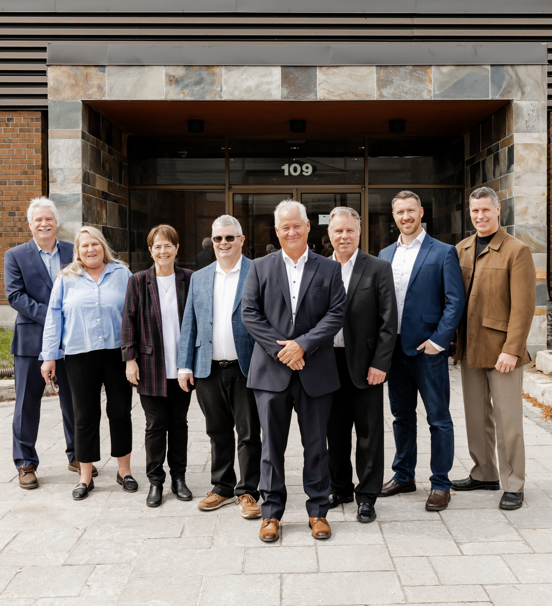 A group portrait of the Lilley Financial team standing outside their office building. From left to right: Mark James, Lois Murphy, Patty Etling, Craig Lilley, Jim Fockens, Philip Rayson, Mike James, and Michael Godfrey. The office building has a brick and stone facade and a sign with '109' above the door.