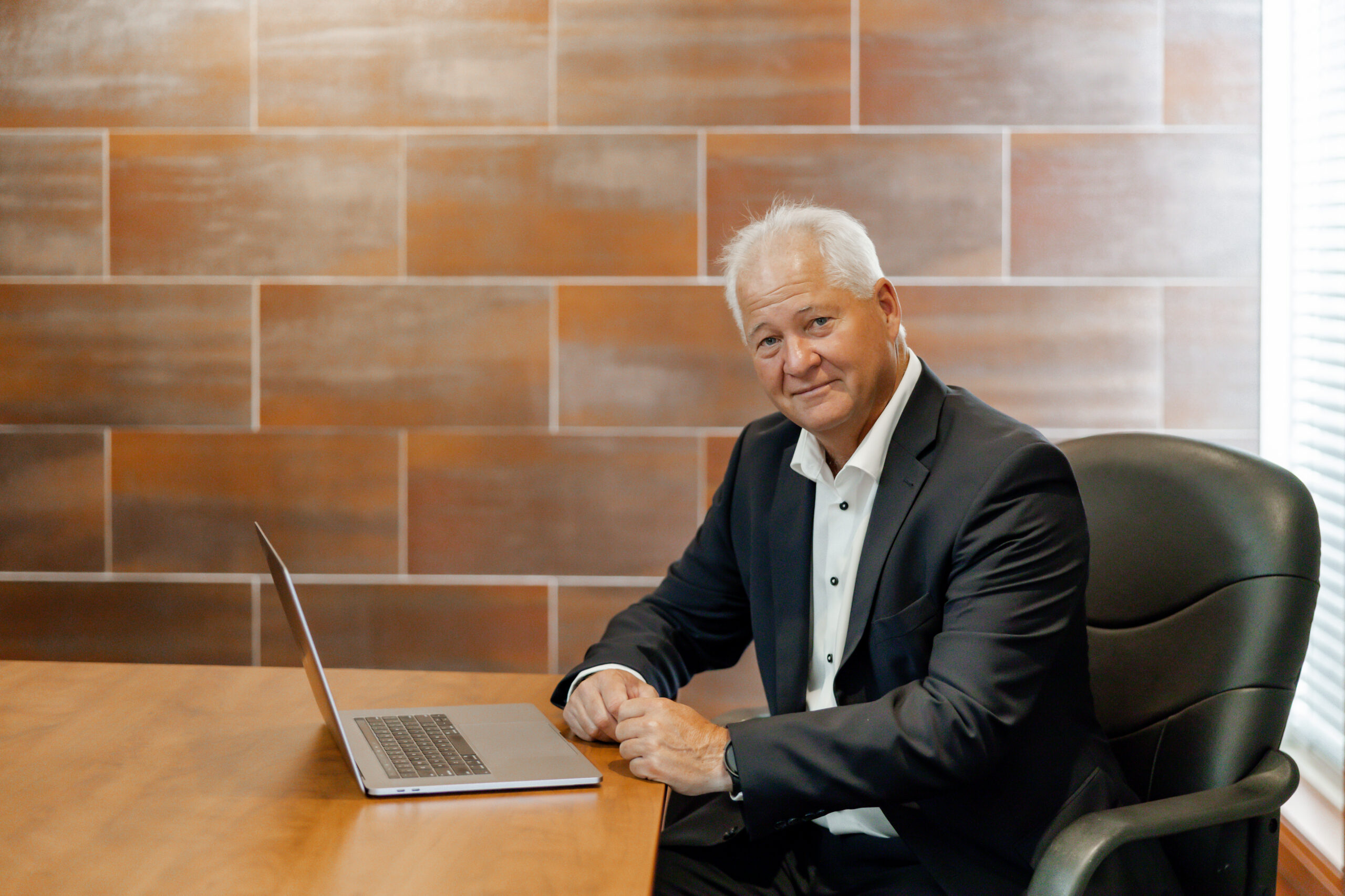 Jim Fockens sits at a wooden conference table with a laptop in front of him, smiling at the camera. He is wearing a dark suit jacket and a white dress shirt. The background is a tiled wall and a window with blinds.