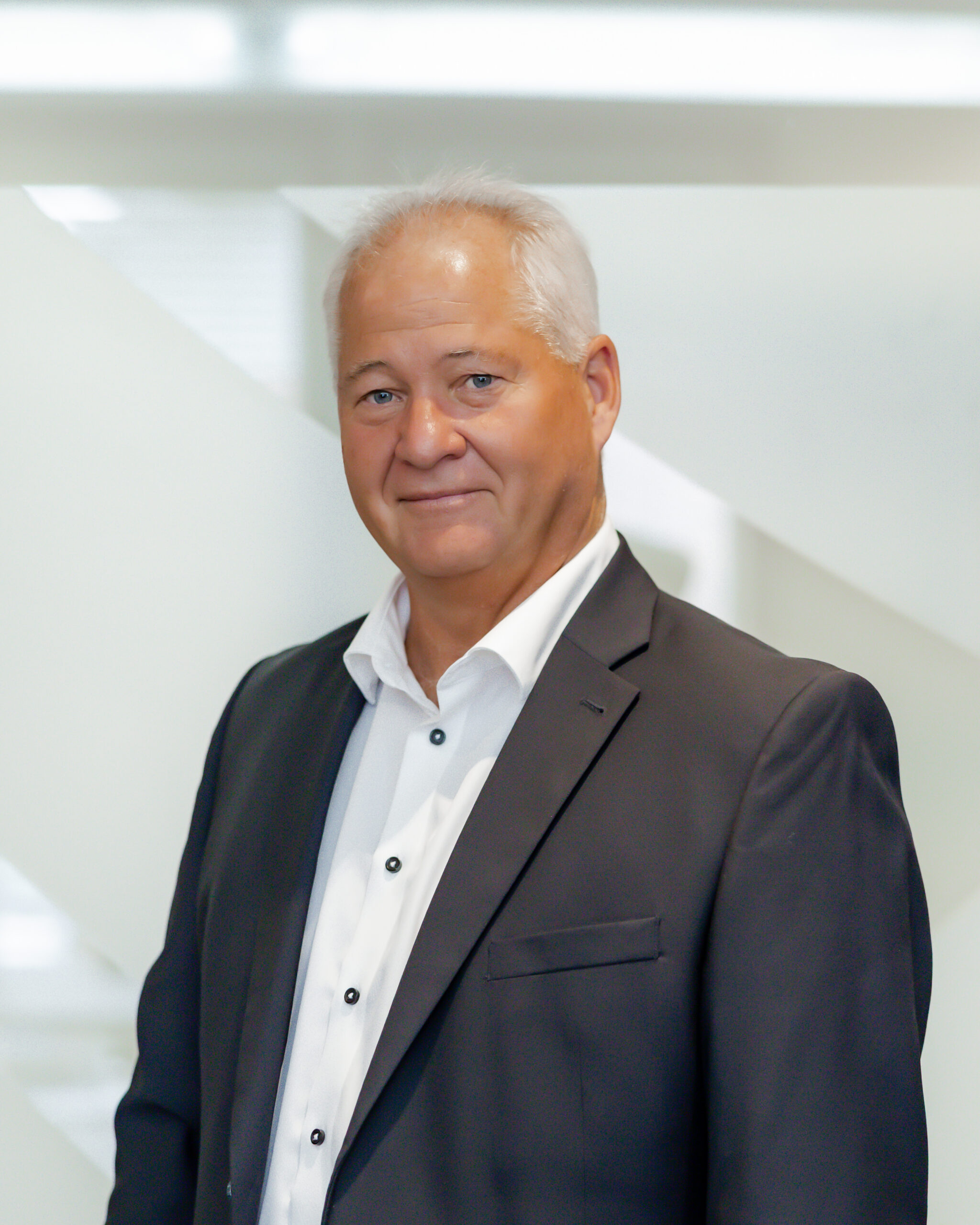A professional portrait of Jim Fockens smiling directly at the camera. He has grey hair, is wearing a dark suit jacket over a white dress shirt, and is standing in front of a modern, white background with geometric shapes.