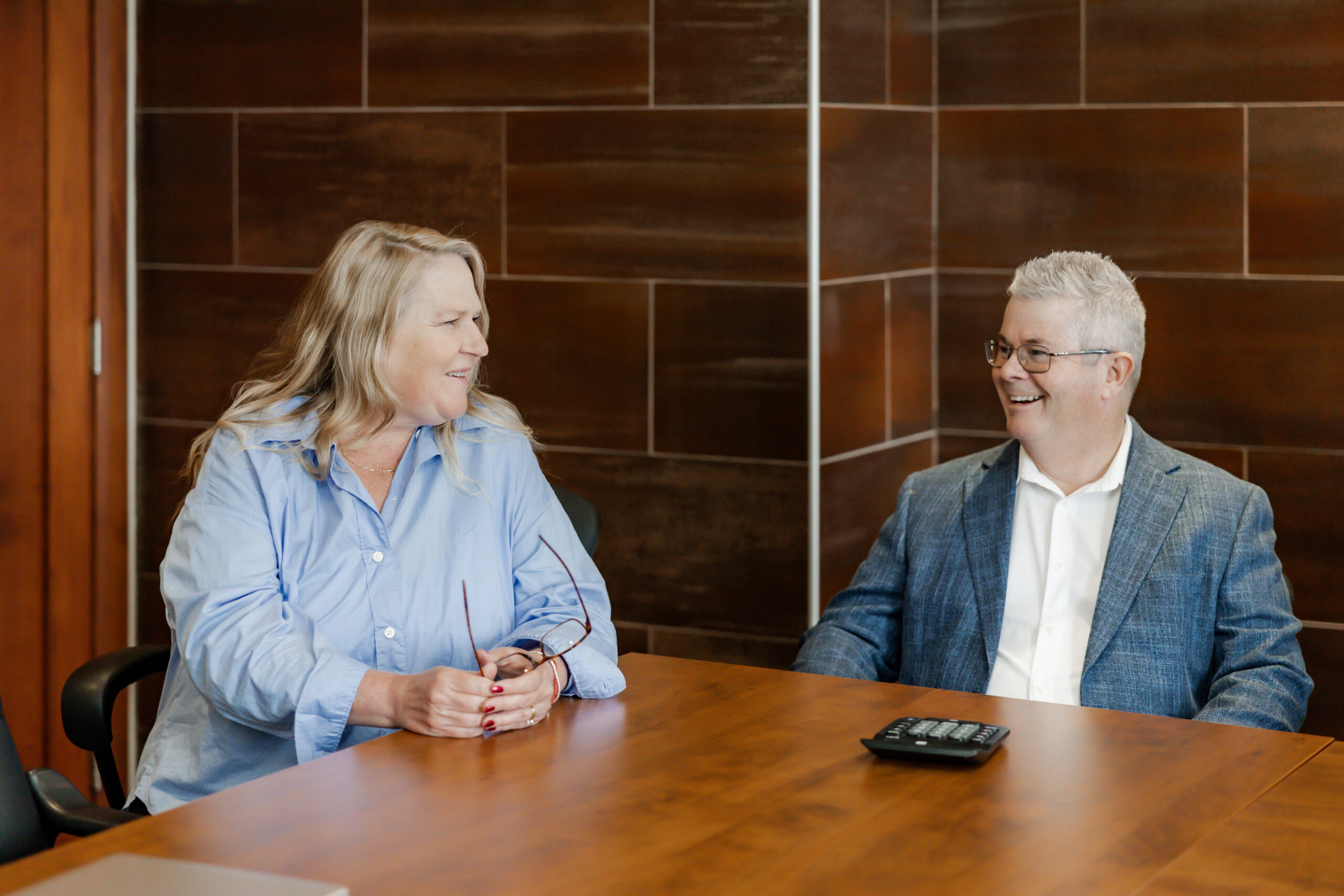 Lois Murphy and Craig Lilley are seated at a wooden conference table, smiling at each other. Lois holds her glasses in her hands and Craig has a calculator on the table in front of him. The background is a tiled wall.