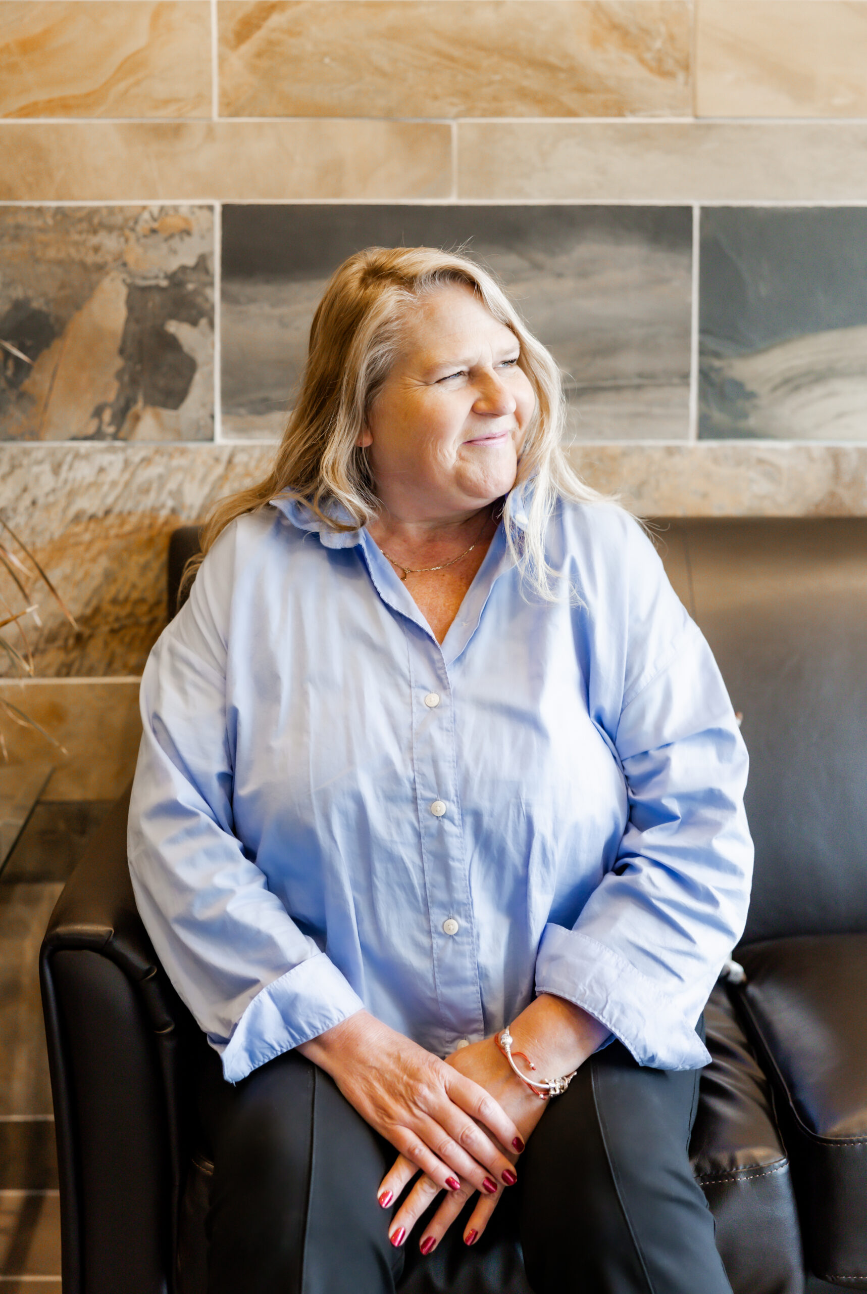 Lois Murphy sits on a black leather couch, smiling and looking to the side. She is wearing a light blue button-up shirt and dark pants, with her hands clasped in her lap. The background is a tiled wall with brown and gray tones.