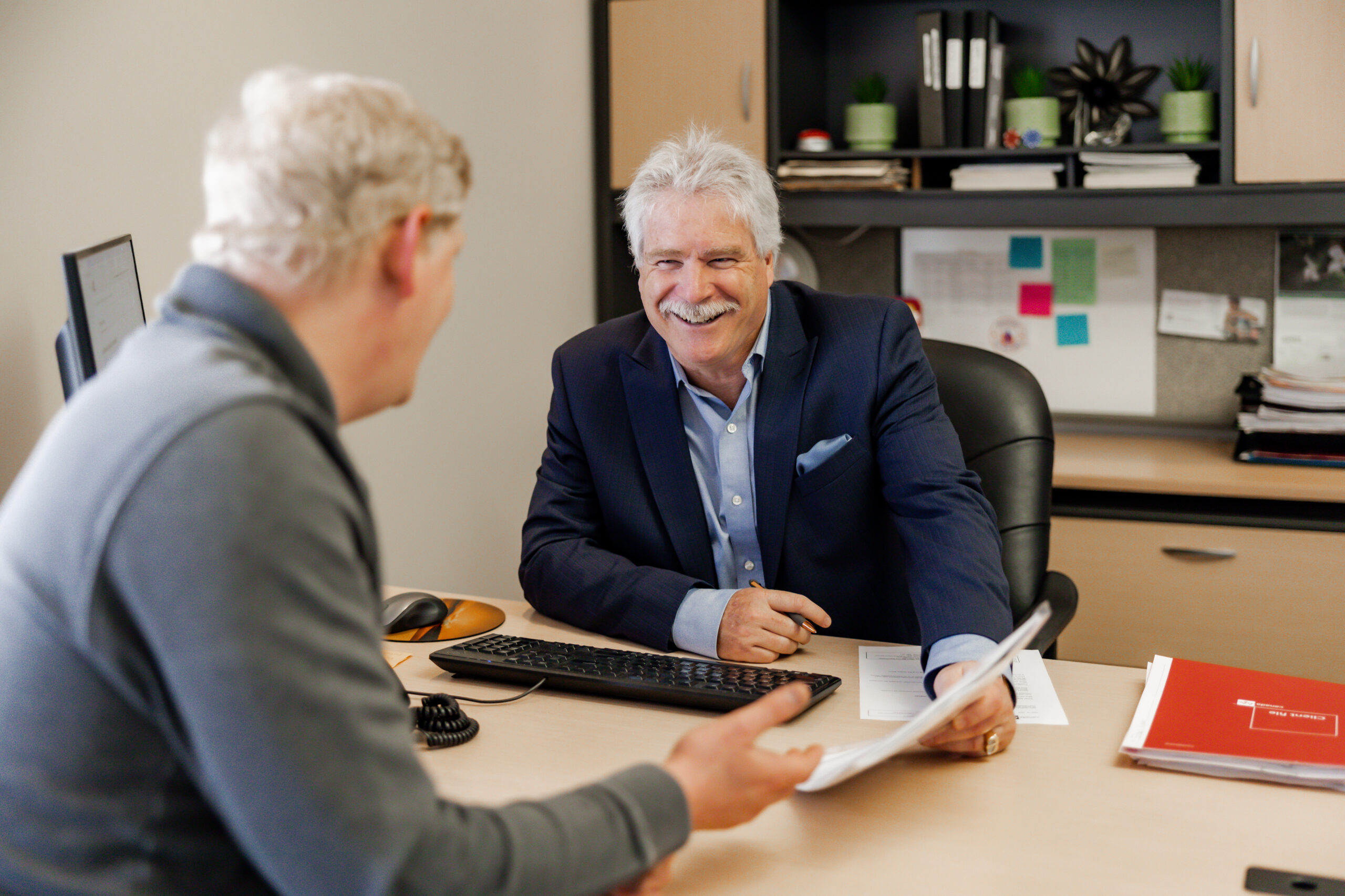 Mark James is seated at his office desk, holding a pen and a document, and smiling while talking with a client who is out of focus in the foreground. Mark is wearing a dark pinstripe suit and is engaged in a friendly conversation. The background shows an office with file organizers and a bulletin board.
