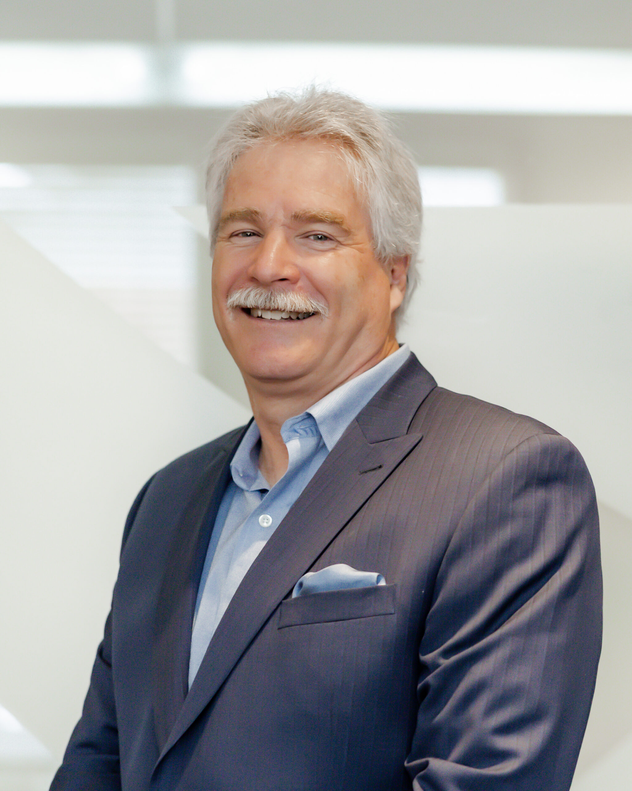A professional portrait of Mark James smiling directly at the camera. He has a mustache, is wearing a dark pinstripe suit, and is standing in front of a modern, white background with geometric shapes.