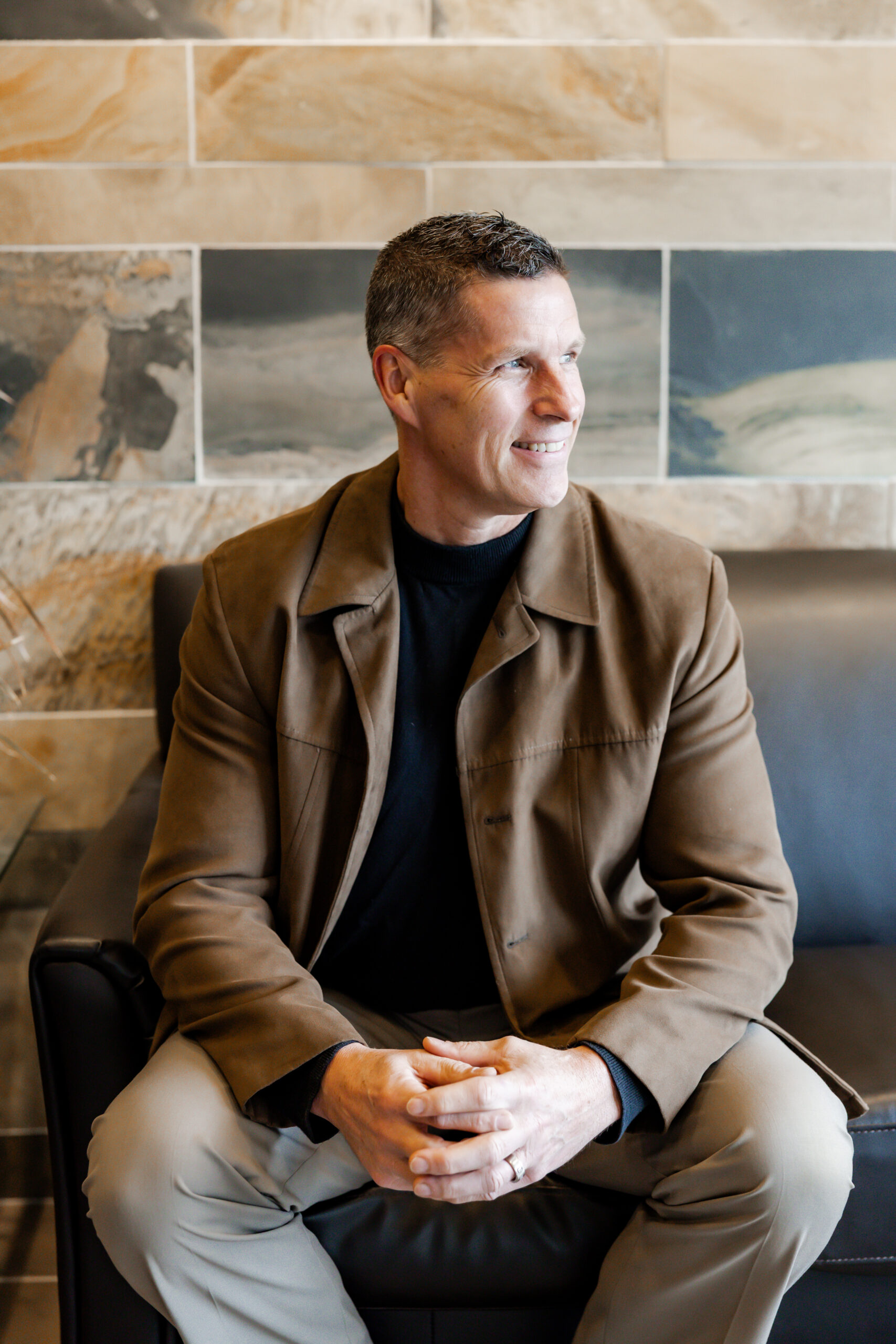 Michael Godfrey sits on a black leather couch, smiling and looking to the side. He is wearing a brown suede jacket over a black shirt and khaki pants, with his hands clasped in his lap. The background is a tiled wall with brown and gray tones.