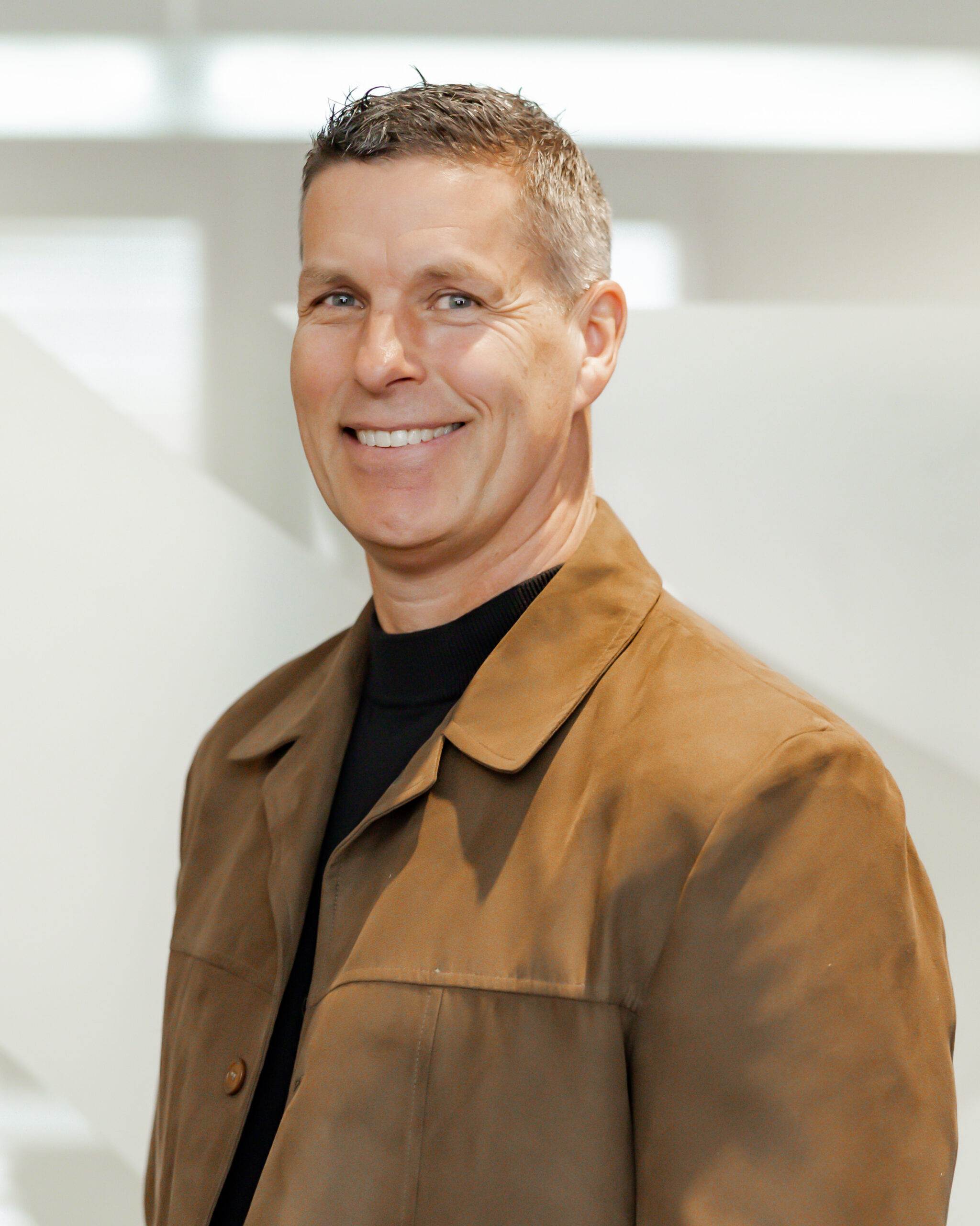 A professional portrait of Michael Godfrey smiling directly at the camera. He is wearing a brown suede jacket over a black shirt, and is standing in front of a modern, white background with geometric shapes.