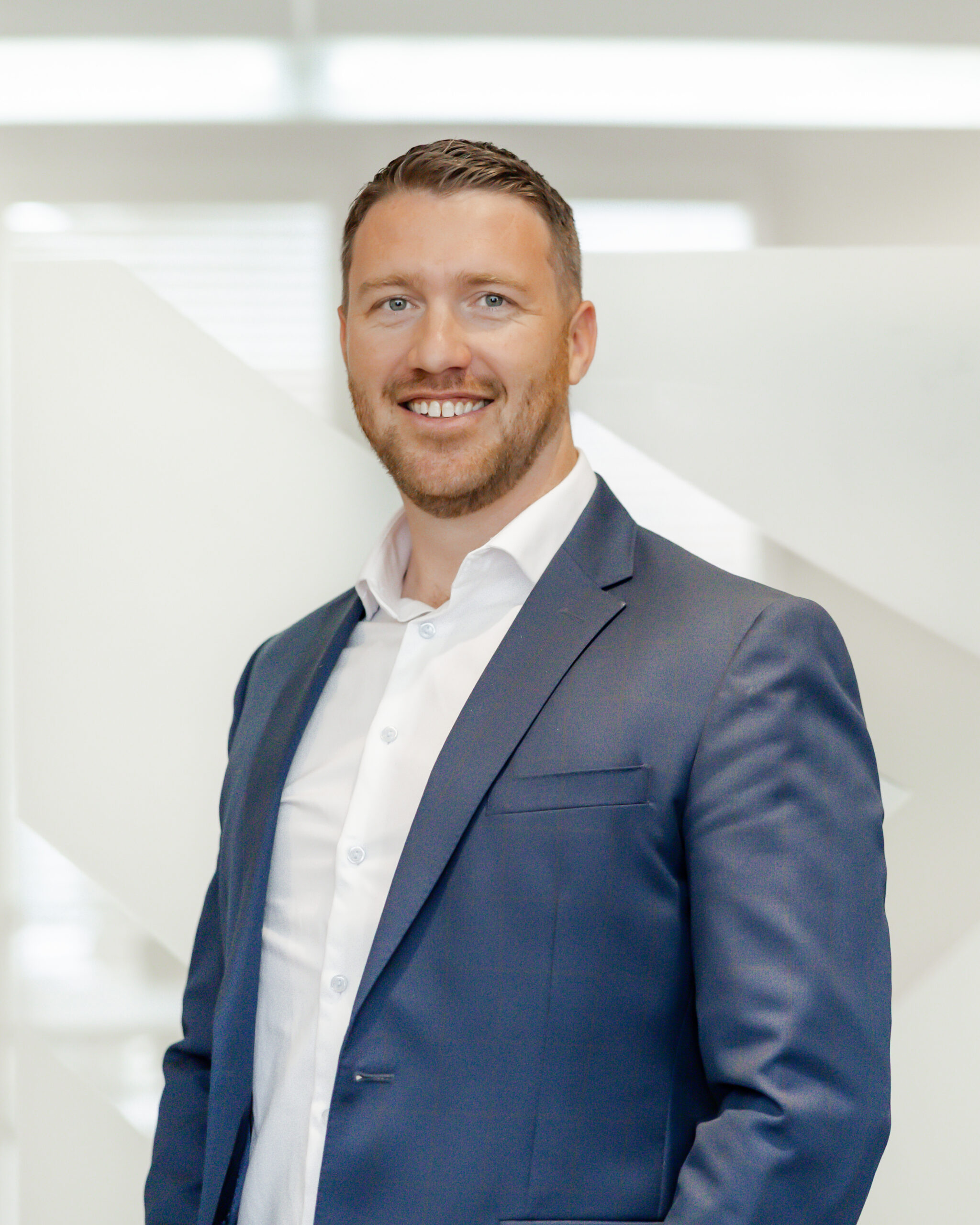 A professional portrait of Mike James smiling directly at the camera. He is wearing a blue suit jacket and a white dress shirt, and is standing in front of a modern, white background with geometric shapes.