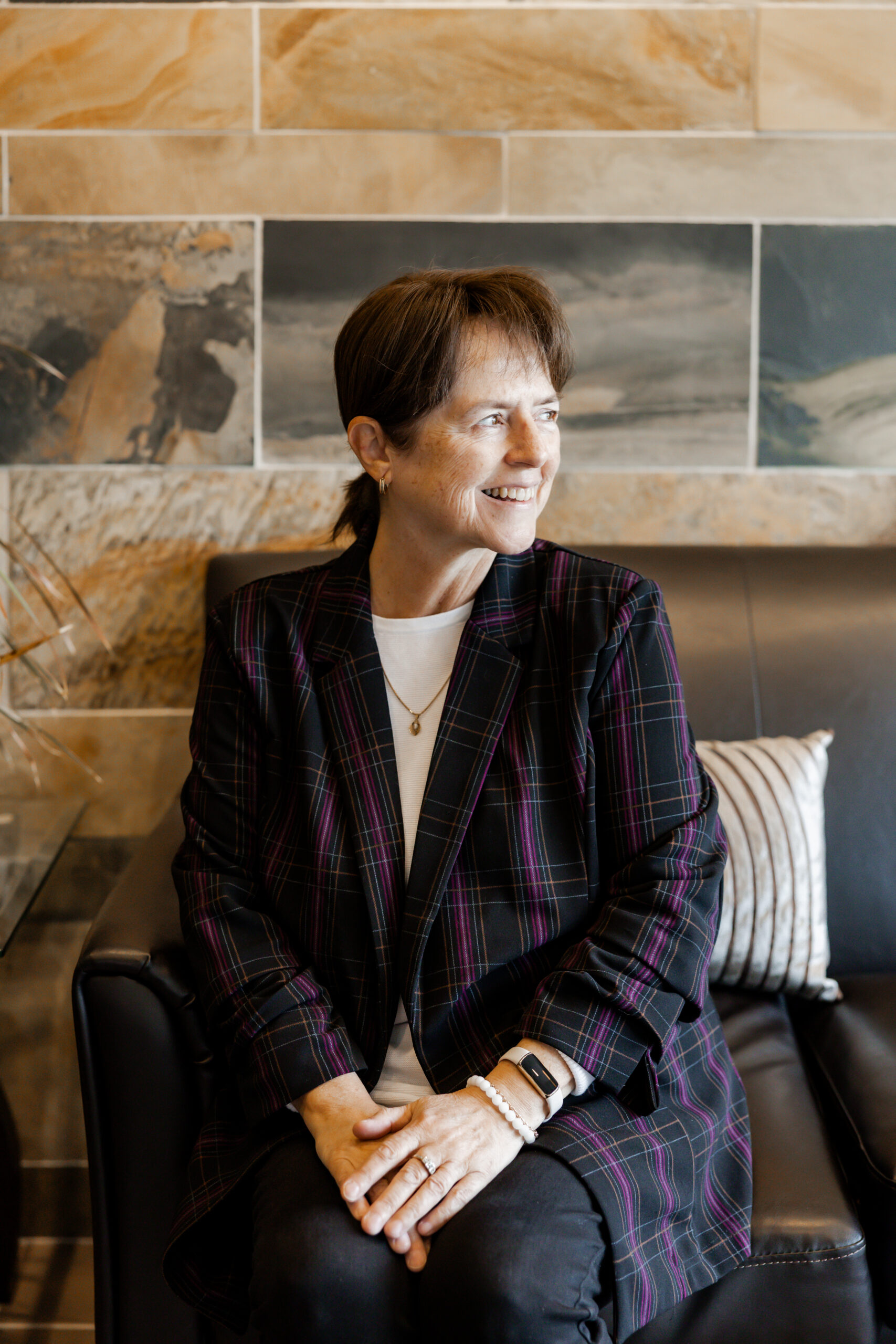 Patty Etling sits on a black leather couch, smiling and looking to the side. She is wearing a dark plaid jacket with purple and red lines over a white shirt, with her hands clasped in her lap. The background is a tiled wall.