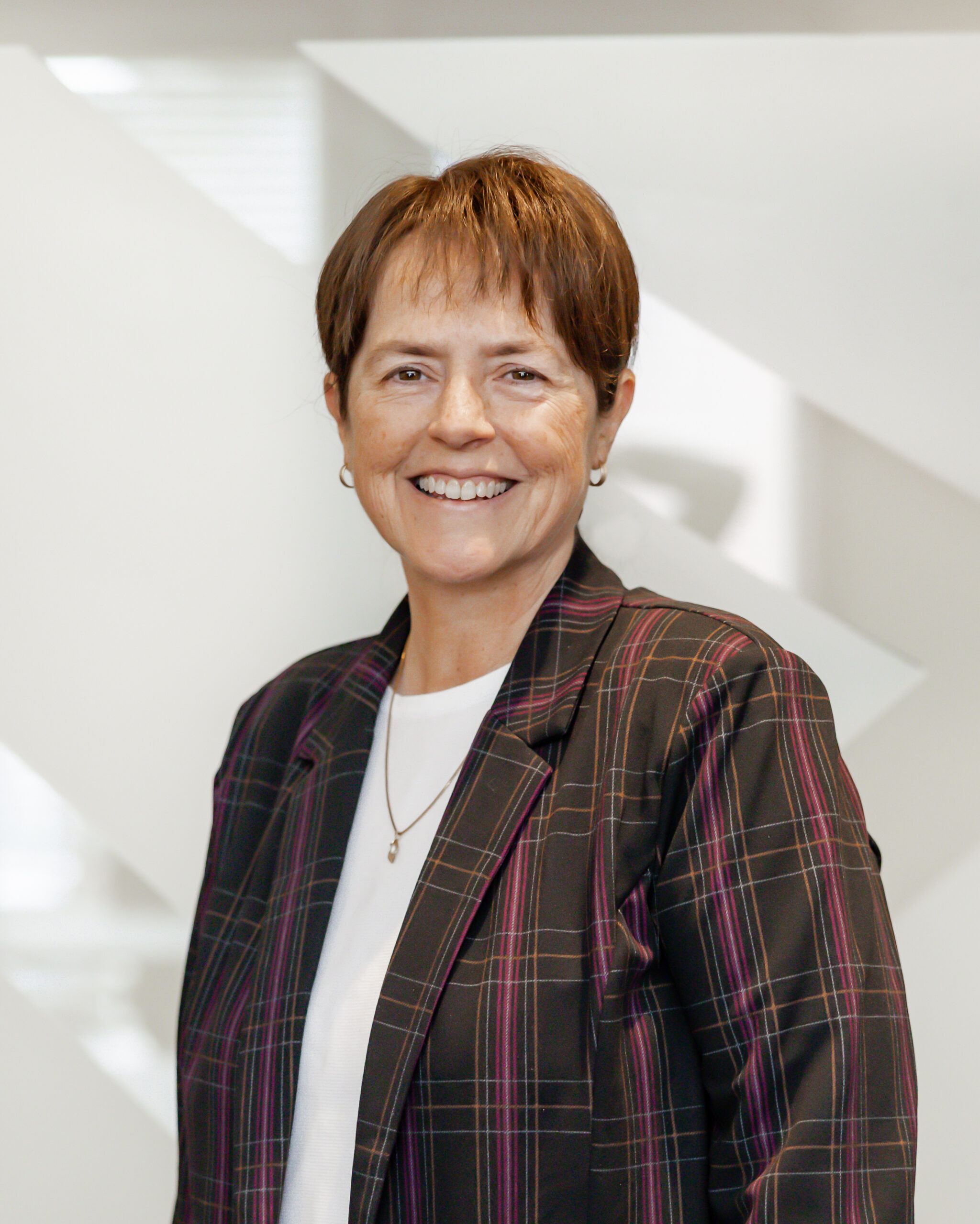 A professional portrait of Patty Etling smiling directly at the camera. She has short brown hair, is wearing a dark plaid jacket over a white shirt, and is standing in front of a modern, white background with geometric shapes.