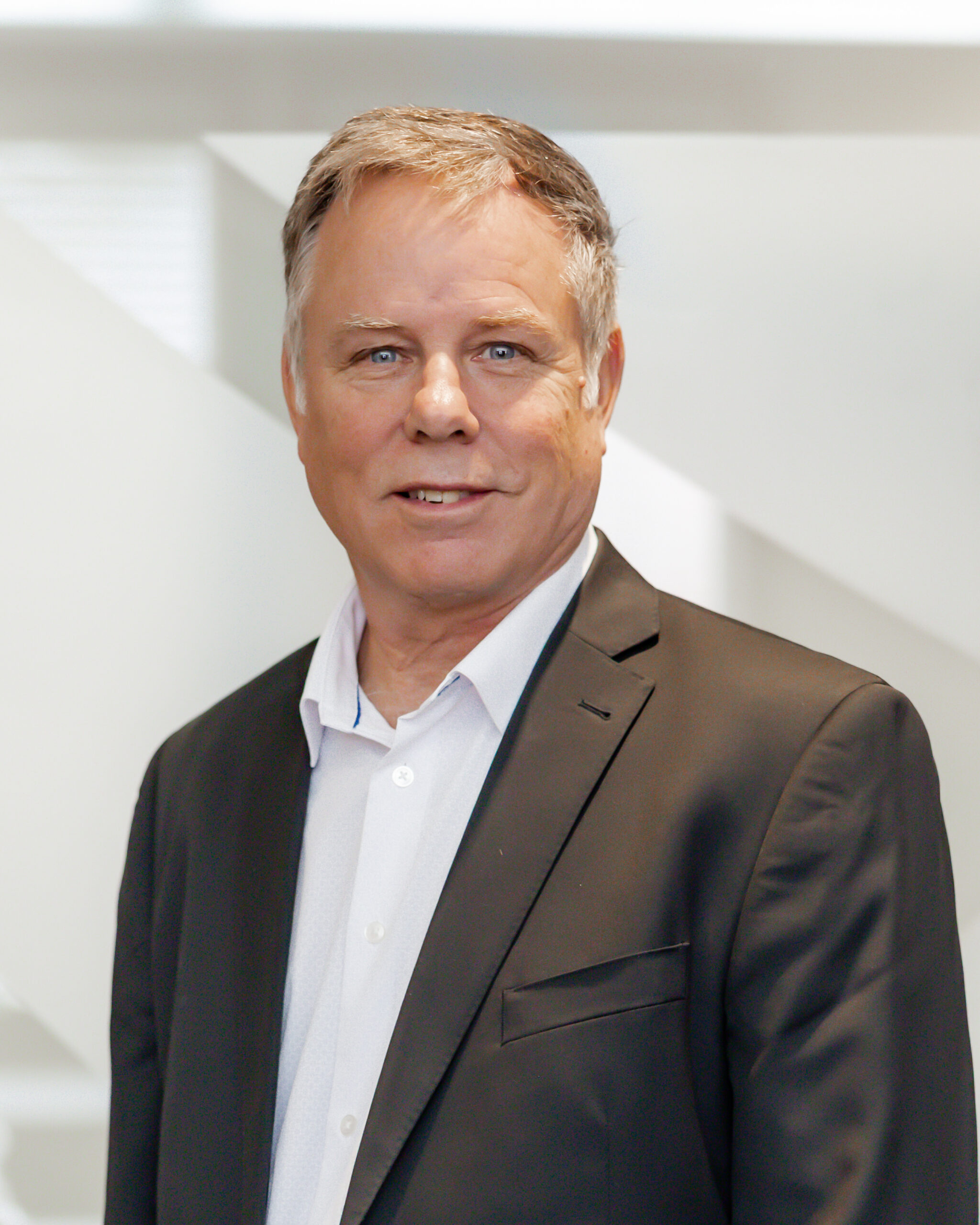 A professional portrait of Philip Rayson smiling directly at the camera. He is wearing a dark suit jacket over a white dress shirt, and is standing in front of a modern, white background with geometric shapes.