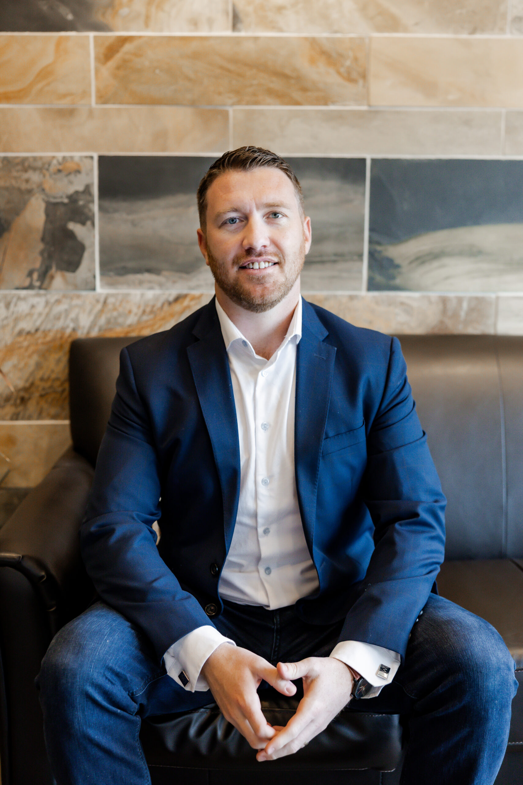 Mike James sits on a black leather couch, smiling directly at the camera. He is wearing a blue suit jacket, a white dress shirt, and jeans, with his hands clasped in his lap. The background is a tiled wall with brown and gray tones.