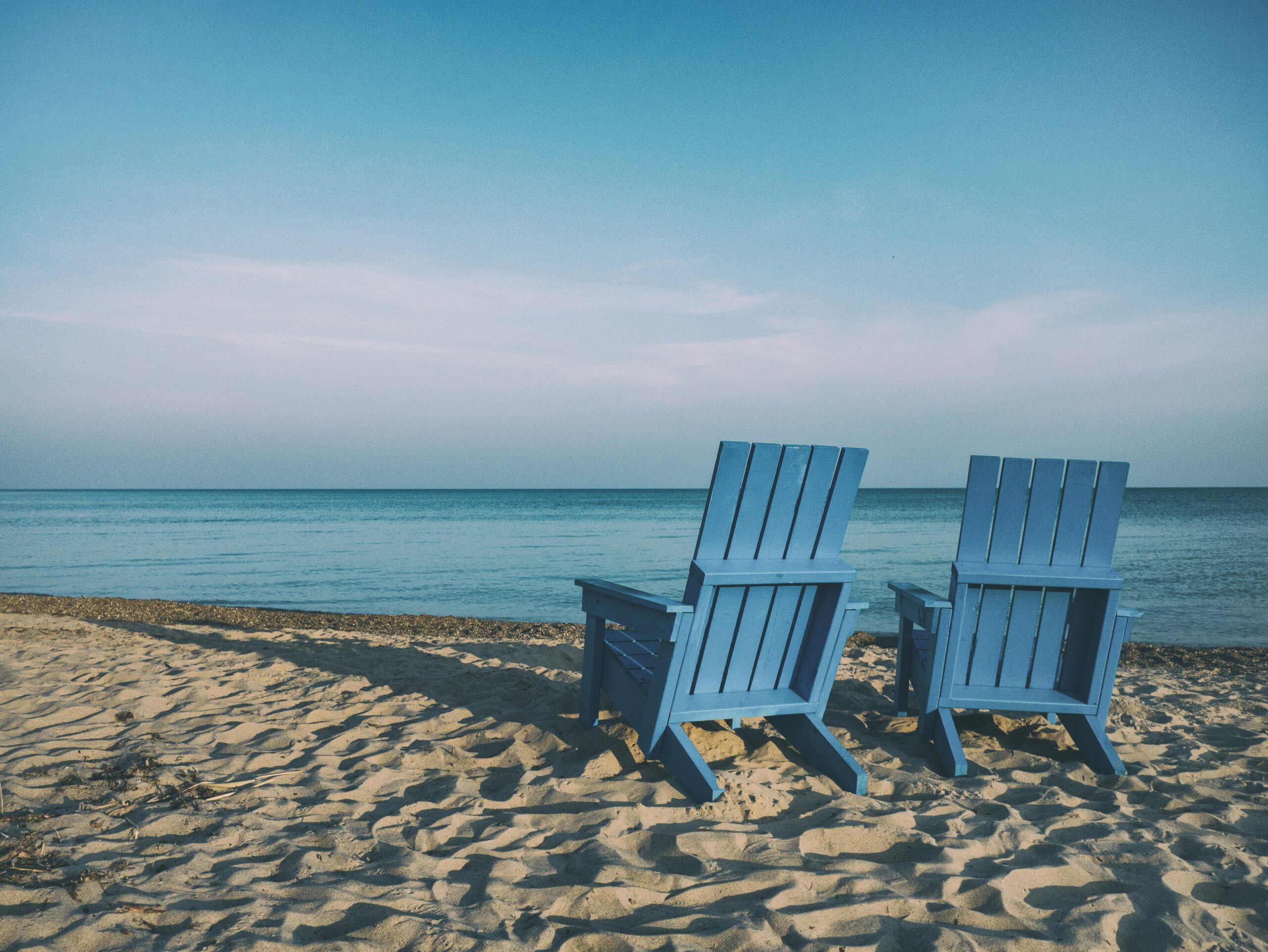 Two blue Adirondack chairs facing a calm lake on a sandy beach under a clear sky.