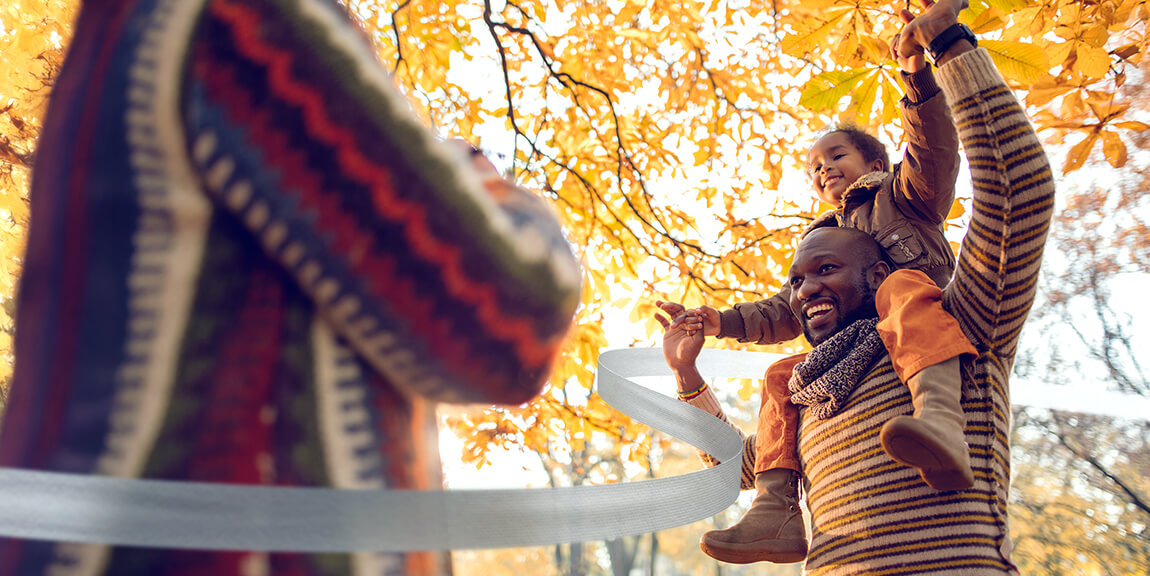 A joyful father carrying his young child on his shoulders in a park filled with golden autumn leaves, both smiling and enjoying the fall weather as another person in a colorful sweater takes their photo.