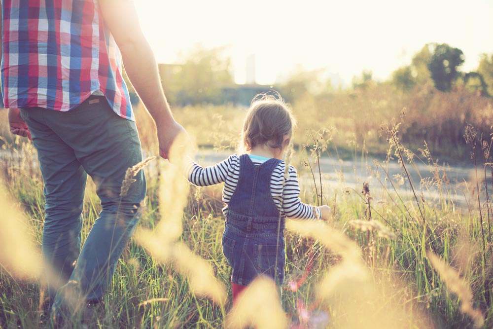 A parent and child walk hand in hand through a sunlit field of tall grass, with the child wearing denim overalls and the adult in a plaid shirt, creating a warm, nostalgic scene in the late afternoon light.